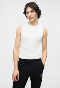 Νεαρό άτομο με μεσαίου μήκους σκούρα μαλλιά, φοράει άσπρο μπλουζάκι χωρίς μανίκια και μαύρο παντελόνι, στέκεται με το ένα χέρι στην τσέπη.