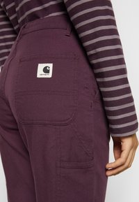 Maroon pants with a back pocket featuring a white label. The wearer is in a striped long-sleeve top, showing the pants' texture and design.