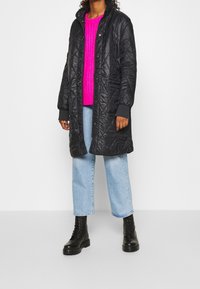 Cappotto lungo trapuntato nero con chiusura a pressione, indossato sopra un maglione lavorato a maglia rosa acceso, abbinato a jeans azzurri chiari e stivali militari neri.
