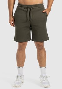 Pantalones cortos de forro polar verde oliva con cinturilla elástica y cordón, que cuentan con bolsillos laterales y un corte relajado, combinados con zapatillas blancas.