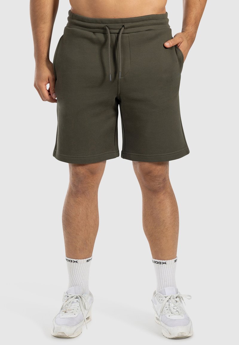 Pantalones cortos de forro polar verde oliva con cinturilla elástica y cordón, que cuentan con bolsillos laterales y un corte relajado, combinados con zapatillas blancas.