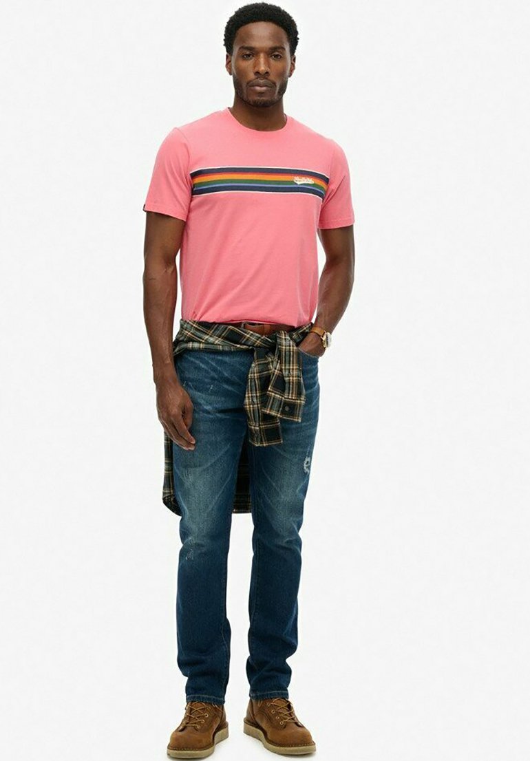 Camisa rosa de manga corta con rayas horizontales coloridas, combinada con pantalones vaqueros azules y botas marrones con cordones; camisa de cuadros atada a la cintura.