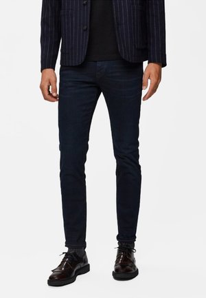 Tmavě modré úzké džíny s hladkým povrchem, v kombinaci s černým tričkem a pinstripovaným sakem. Celý outfit doplňují kožené boty.