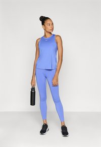 Αθλητική φανέλα σε ανοιχτό γαλάζιο και κολάν, εφαρμοστά και με απαλό ύφασμα. Μαύρο παγούρι νερού στο ένα χέρι, μαύρα αθλητικά παπούτσια. Απλό φόντο.