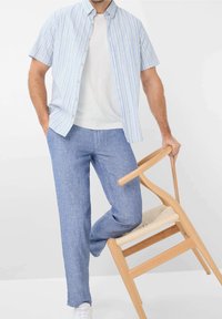 Outfit da uomo con una camicia a maniche corte a righe blu chiaro e pantaloni blu chiaro texture, abbinati a sneakers bianche e una sedia di legno.