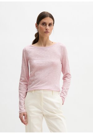 Frau mit braunen Haaren im Pferdeschwanz, die ein hellrosa Langarmshirt und cremefarbene Hosen trägt und vor einem schlichten weißen Hintergrund steht.