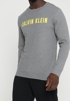 Mann trägt ein graues Langarmshirt mit gelbem "CALVIN KLEIN"-Schriftzug auf der Brust und schwarze Hose vor einfarbigem Hintergrund.
