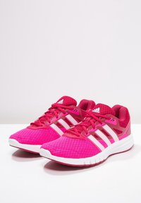 Scarpe da ginnastica rosa con tomaia in rete, design a tre strisce bianche e suola testurizzata. Presentano un colletto imbottito e un'imbottitura leggera.