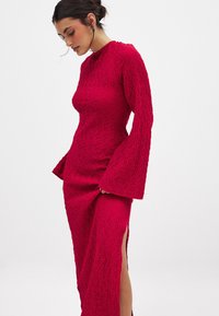 Red textured dress with long bell sleeves, knee-high slits on both sides, and a fitted silhouette, made from a woven fabric.