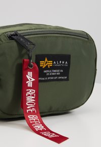 Borsetta in nylon verde oliva con cerniera nera, dotata di un'etichetta nera e un'etichetta in tessuto rossa con la scritta "RIMUOVERE PRIMA DEL Volo."