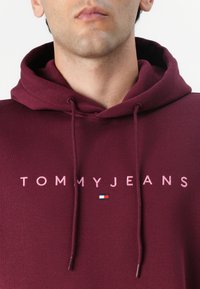 Burgunderfarget hettegenser med snøring og stor "TOMMYJEANS"-logo i rosa; har et lite brodert flaggemblem i rødt, hvitt og blått.