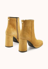Bottes cheville en daim camel avec un bout carré, un talon bloc haut et une fermeture éclair latérale. Présente une texture lisse et des accents matériels minimaux.