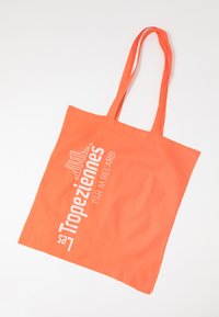 Borsa in cotone arancione con due manici lunghi. Presenta testo stampato in bianco: "Les Tropéziennes par M. Belarbi." Texture liscia, forma rettangolare.