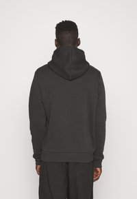 Man die met de rug naar de camera staat, draagt een zwarte hoodie met de capuchon naar beneden en een zwarte broek, tegen een effen achtergrond.