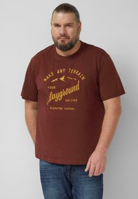 Ciemnobrązowy t-shirt z żółtym napisem i grafiką, krótki rękaw, okrągły dekolt. Nosi się go z niebieskimi dżinsami.