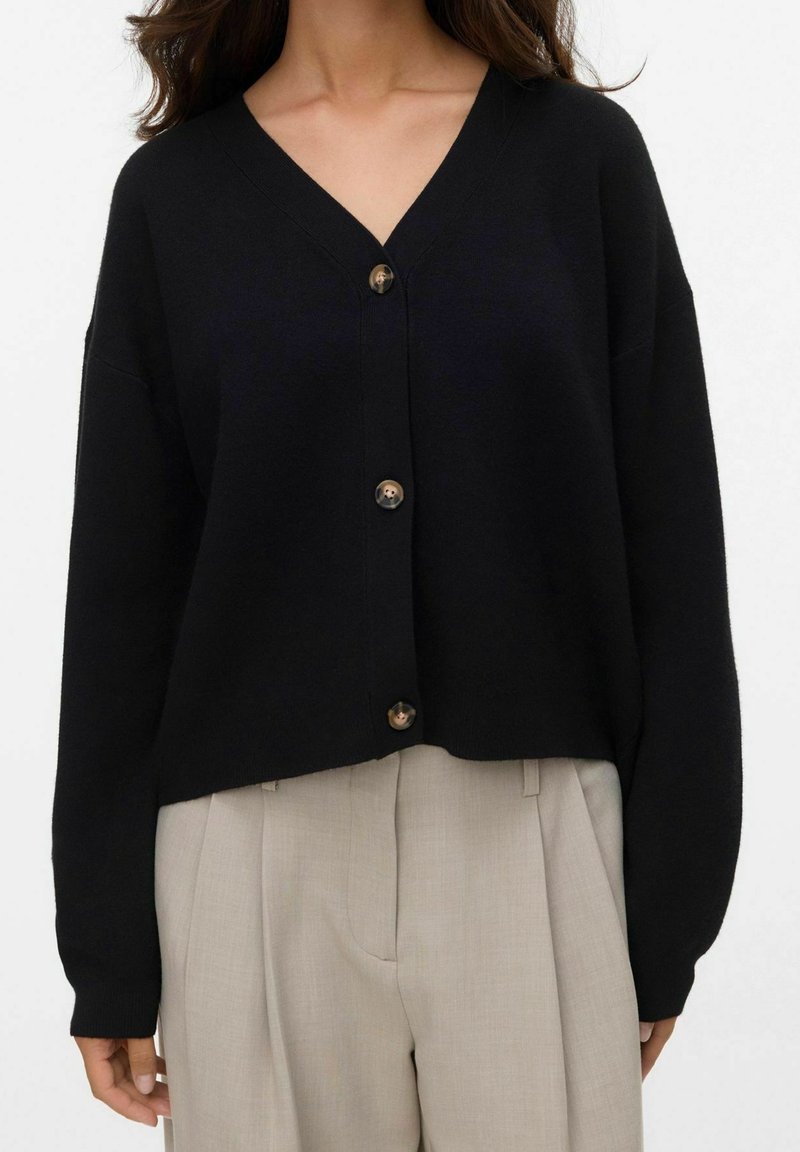 Femme portant un cardigan noir à boutons col en V et un pantalon plissé beige, vue du cou jusqu'aux hanches sur fond blanc.