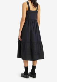 Robe en coton noire à design à volants, bretelles ornées de dentelle, coupe mi-longue, associée à des chaussettes foncées et des chaussures noires épaisses.