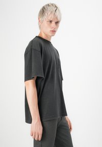 Tricou supradimensionat de culoare gri închis, realizat din bumbac, cu mâneci scurte și un buzunar pe partea stângă. Textură netedă cu detalii de cusătură vizibile.