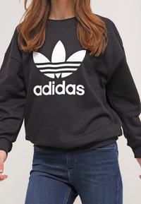 Svart sweatshirt med stor vit Adidas-logotyp, med klassisk rundad halsringning och långa ärmar, tillverkad av ett mjukt tyg.