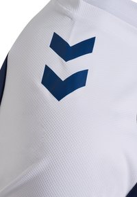 Biała sportowa koszulka z teksturowaną tkaniną, z granatowymi wzorami w kształcie szewronów po prawej stronie. Rękawy mają dopasowany krój.