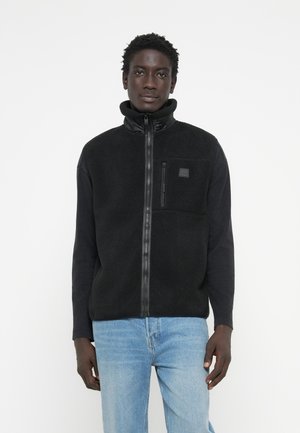 Black fleece jacket with a high collar, zip front, and a small zippered chest pocket. Soft texture, paired with light blue jeans.