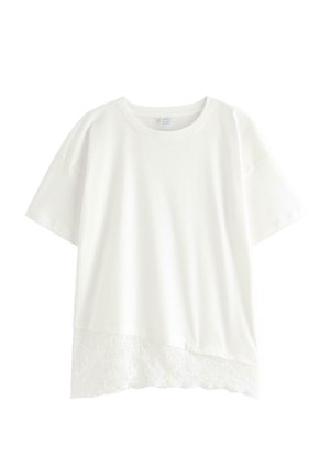Jonge vrouw staat glimlachend, draagt een wit T-shirt met korte mouwen over een kanten top, lichtblauwe spijkerbroek, roze sneakers en draagt een kleine zwarte tas.