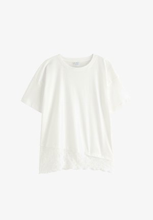 Eine junge Frau steht lächelnd da, trägt ein weißes Kurzarmshirt über einem Spitzenoberteil, hellblaue Jeans, pinke Turnschuhe und trägt eine kleine schwarze Tasche.