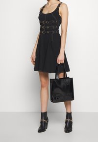 Kobieta w czarnej sukience bez rękawów z trzema zapięciami na klamry oraz czarnych botkach za kostkę, trzymająca torbę typu tote od Versace Jeans Couture.