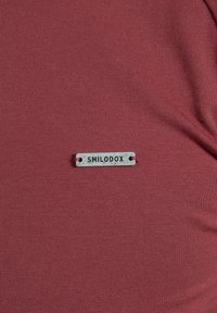 Närbild av ett vinrött tyg med en liten rektangulär silveretikett som har texten "SMILODOX" sydd på.