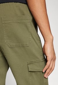 Olivově zelené cargo kalhoty s kapsou na nášivku, prošívanými detaily a hladkou texturou, s detailním pohledem na pravou stranu.
