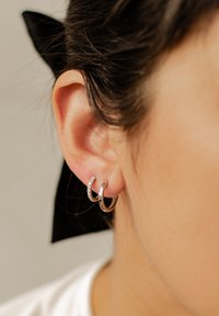 Srebrne kolczyki w kształcie obręczy z gładkim wykończeniem i rzędem małych, przezroczystych kryształków po jednej stronie, noszone na uchu modelki.