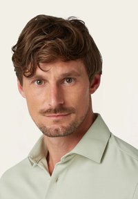 Mannelijke model draagt een lichtgroene polo met een kraag en een enkele knoopdetail, die een gladde textuur en een op maat gemaakte pasvorm laat zien.