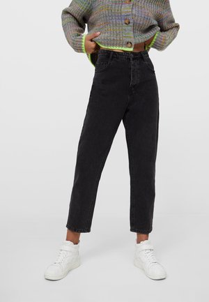 Personne portant un jean noir taille haute, des baskets blanches et un cardigan court multicolore avec des boutons en bois sur un fond uni.
