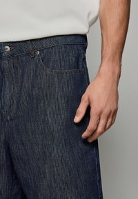 Jeans din denim de culoare albastru închis cu un model de dungi verticale, inclusiv buzunare frontale, sistem de închidere cu nasturi și detalii de cusătură vizibile.