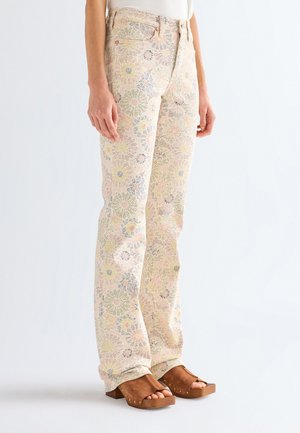 Kvinde iført cremefarvede bukser med høj talje og pastelblomstret mønster samt brune platformsandaler med åbne tæer, stående mod en ensartet baggrund.