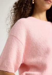 Kvinde med krøllet hår iført en blød, lyserød, kortærmet striktrøje mod en ensfarvet baggrund.