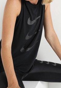 Czarny bezrękawnik sportowy z gładkiego materiału, okrągłym dekoltem i wieloma błyszczącymi logo Nike Swoosh na przodzie.