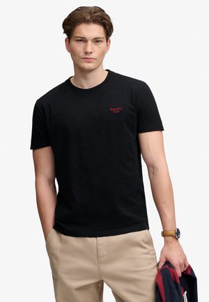Młody mężczyzna w czarnej koszulce Superdry i beżowych spodniach, trzymający w lewej ręce złożoną czerwoną i granatową kurtkę.