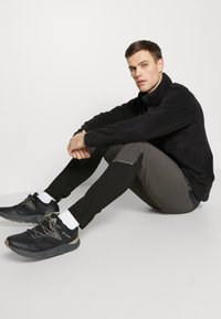 Black fleece jacket, gray and black athletic pants, black shoes with gray accents, sitting posture, plain background.