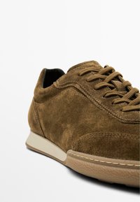 Sneaker in suede marrone con texture liscia, design con lacci, suola di colore chiaro e dettagli di cucitura discreti. Inquadratura ravvicinata che mette in risalto il materiale.