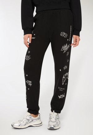 Persona che indossa pantaloni jogger neri con testo grafico e simboli bianchi, abbinati a scarpe da ginnastica argentate e bianche.