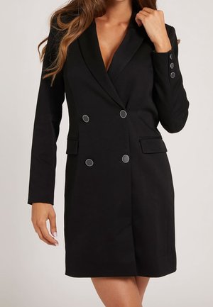 Frau trägt ein schwarzes doppelt geknöpftes Blazer-Kleid mit silbernen Knöpfen, langen Ärmeln und Klappentaschen, vor einem schlichten hellen Hintergrund.