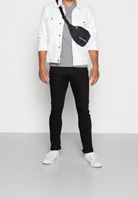 Man som bär en vit denimjacka, grå skjorta, svarta jeans, vita sneakers och en svart axelväska framför en enfärgad vit bakgrund.