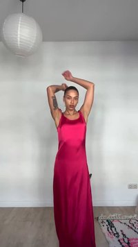 Γυναίκα με αμάνικο κόκκινο σατέν φόρεμα σηκώνοντας τα χέρια πάνω από το κεφάλι της, στέκεται σε ένα λιτό δωμάτιο με λευκό χαρτονένιο φανάρι και χαλί με σχέδια.