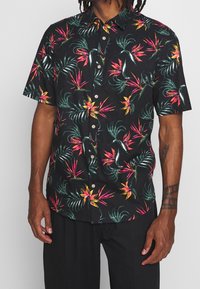 Korte mouwen zwarte shirt met een levendige tropische print van roze en groene bladeren en bloemen. Knopsluiting aan de voorkant met een kraag.