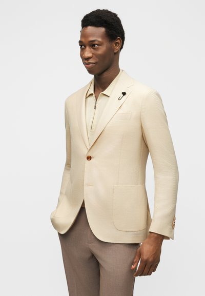 Άνδρας που φοράει κρεμ σακάκι με ένα κουμπί, μπεζ πουκάμισο με φερμουάρ και καφέ παντελόνι, στέκεται με το ένα χέρι στην τσέπη σε λευκό φόντο.
