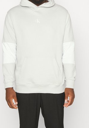 Hombre vestido con una sudadera gris clara con paneles de manga blanca y bolsillo frontal, combinada con pantalones negros, frente a un fondo sencillo.