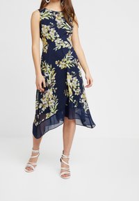 Marineblauw bloemenjurk met een vloeiende uitstraling, voorzien van gele en groene bloemmotieven. Het materiaal lijkt lichtgewicht met een hi-low zoomlijn.