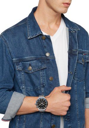 Montre chronographe argentée avec cadran noir, accents bleus, et bracelet en acier inoxydable. Portée au poignet, associée à une veste en denim bleue.