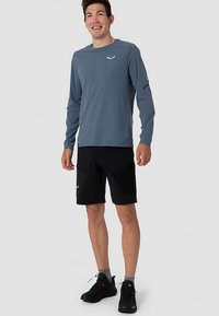 Fiatal férfi áll, hosszú ujjú kék sportpólót, fekete nadrágot, szürke zoknit és fekete futócipőt visel, enyhén mosolyog.